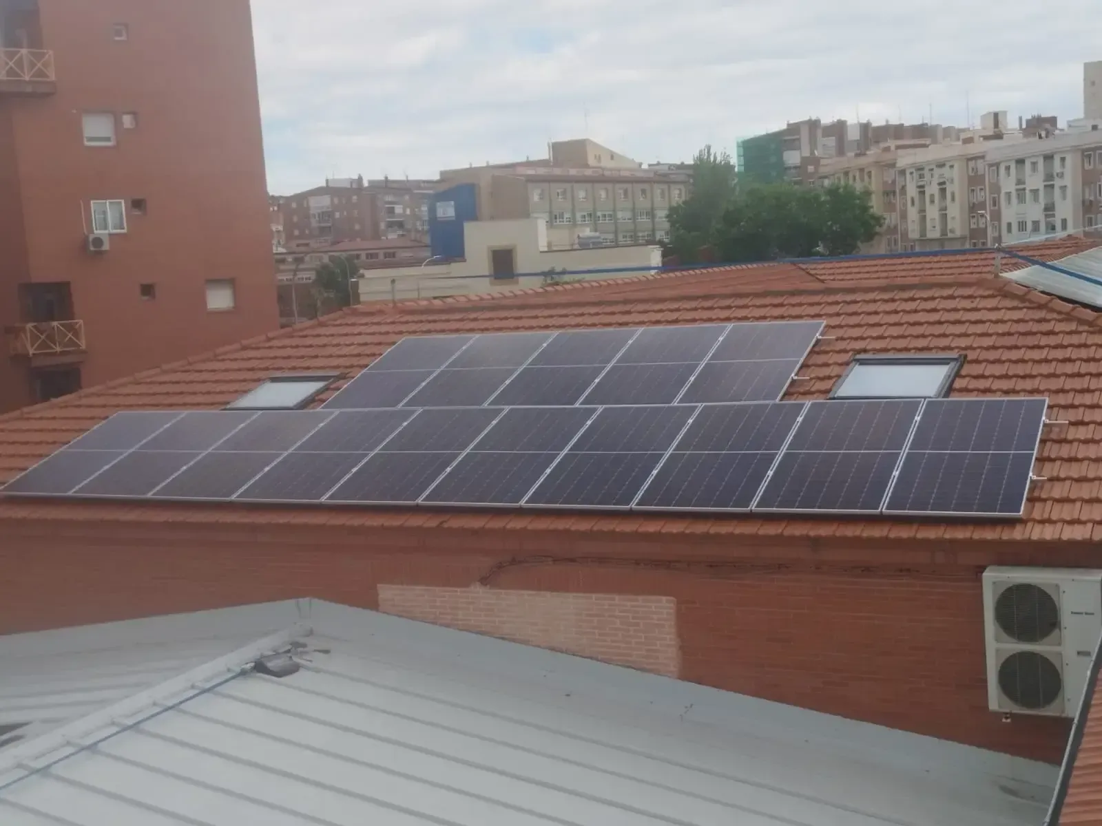 Paneles solares en la azotea de un edificio con tejas rojas y edificios de la ciudad al fondo.