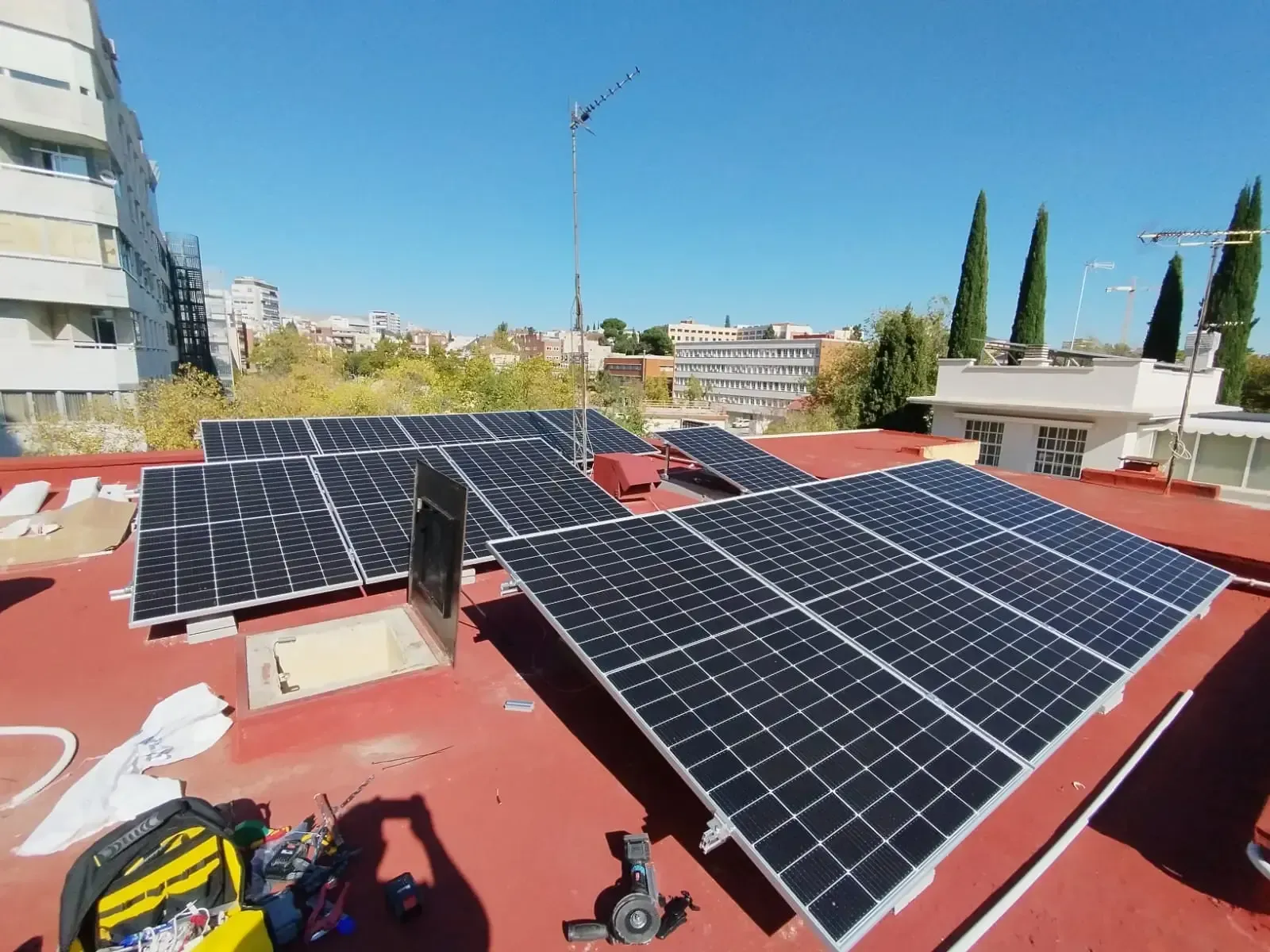 Paneles solares en un tejado rojo con herramientas cerca y edificios a lo lejos bajo un cielo azul despejado.