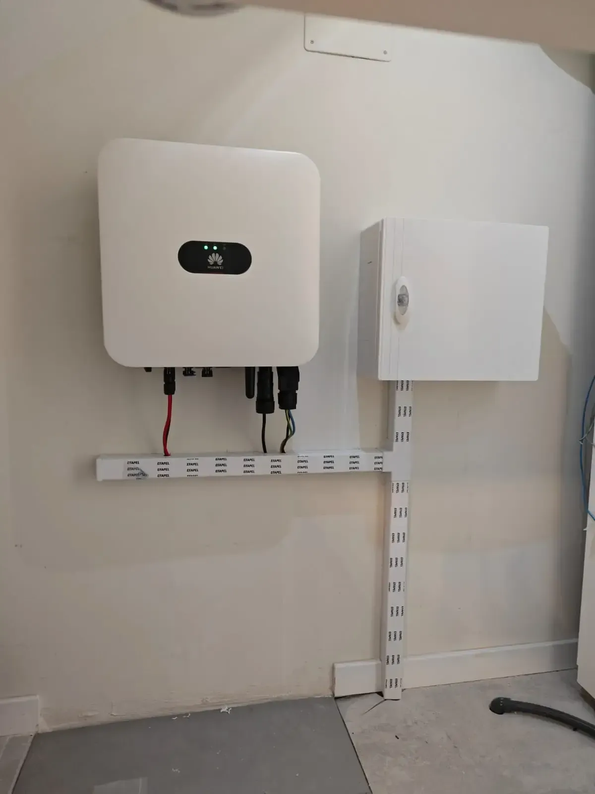 Inversor eléctrico mural y caja de control blanca con cableado a la vista en una pared interior alicatada.