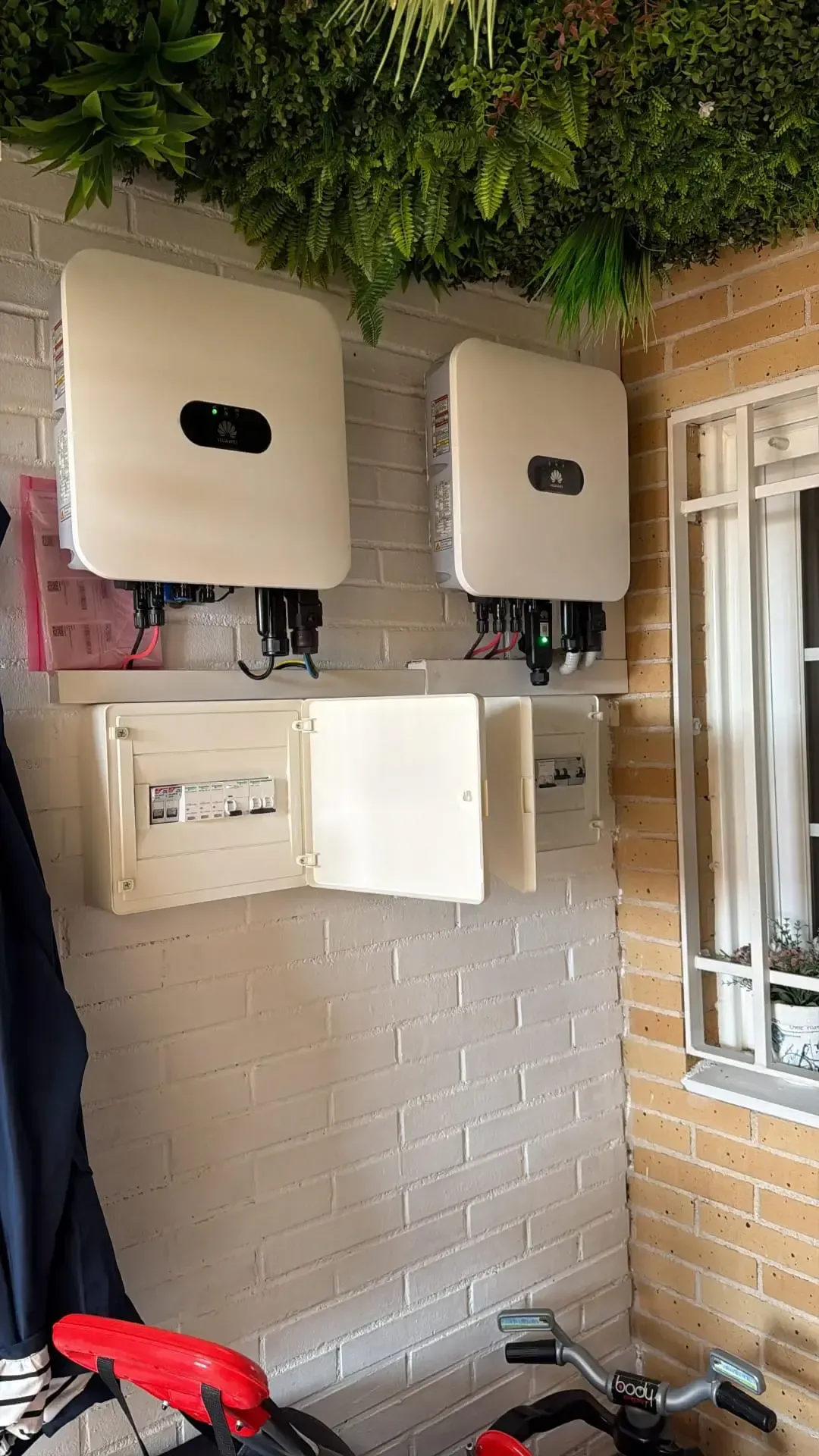 Dos inversores solares montados en la pared sobre un panel eléctrico blanco en un patio de ladrillo.