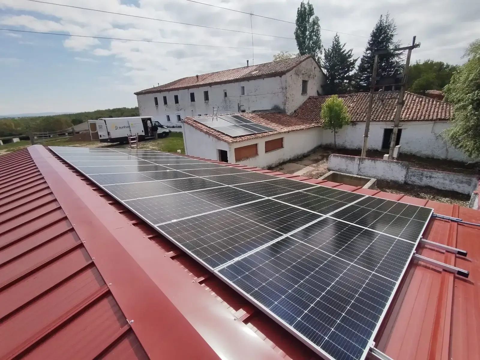 Paneles solares instalados en un techo de metal rojo con vistas a un edificio rural y árboles.
