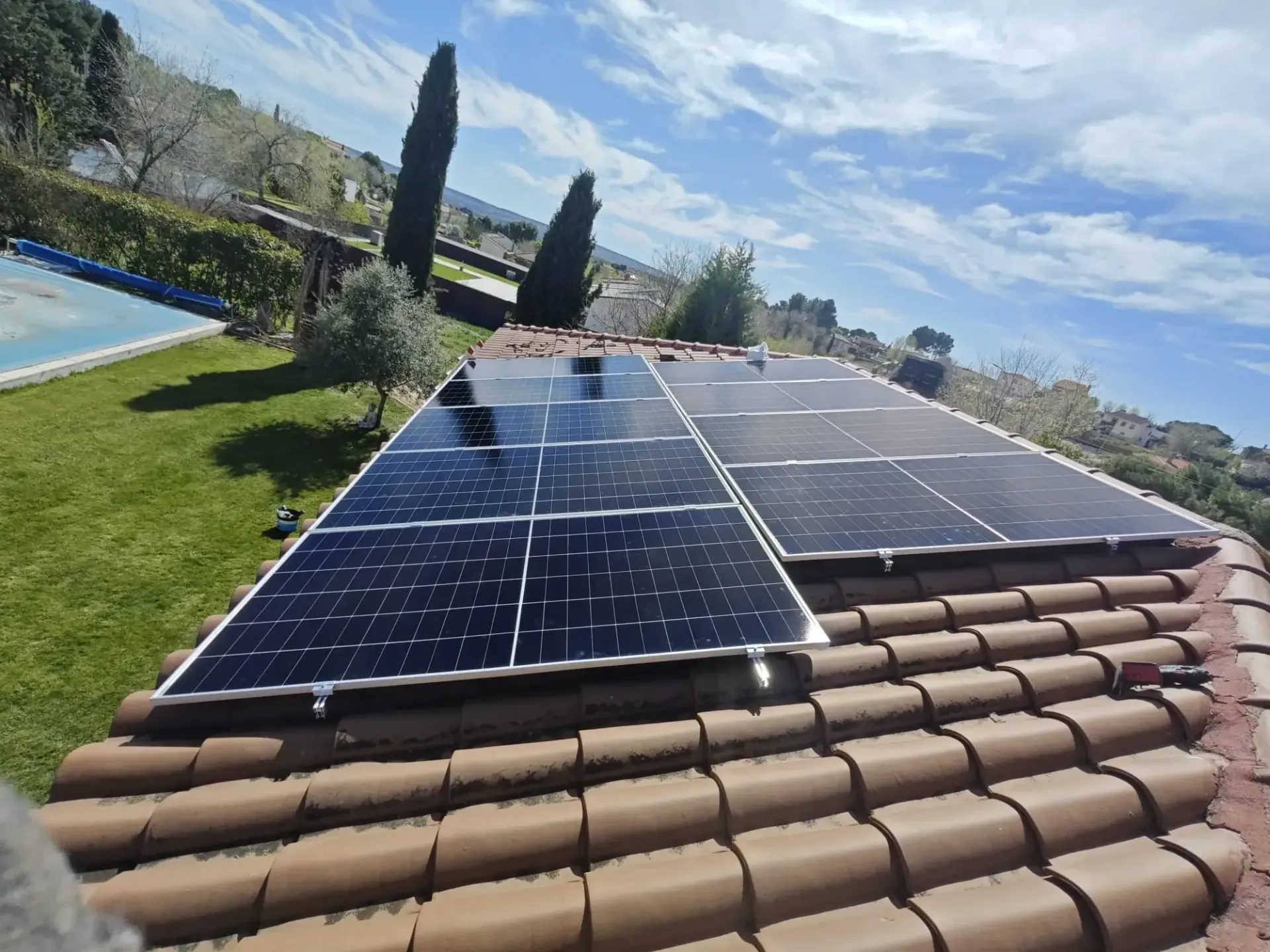 Paneles solares instalados en un tejado de tejas de terracota junto a una piscina y un césped verde bajo un cielo azul.