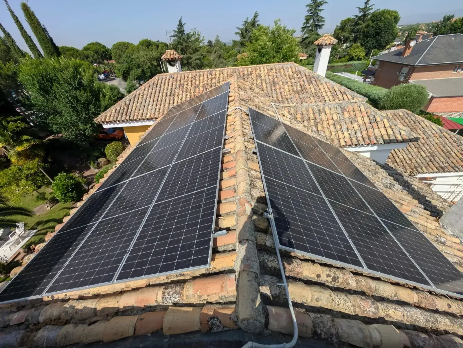 Paneles solares instalados en un tejado de tejas, con árboles y casas vecinas al fondo.