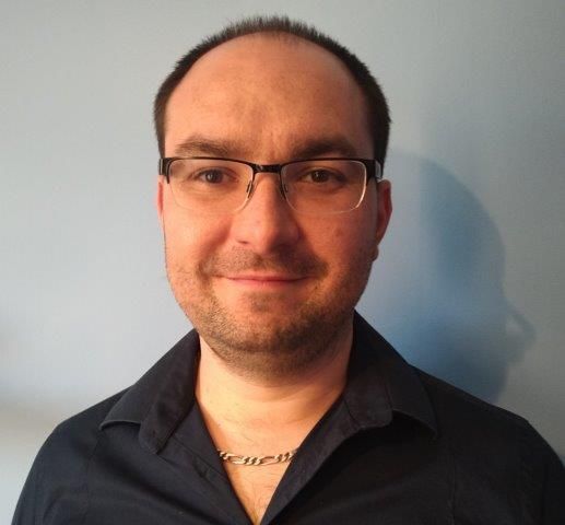 Portrait d'une personne souriante portant des lunettes et une chemise foncée, sur un mur bleu clair.