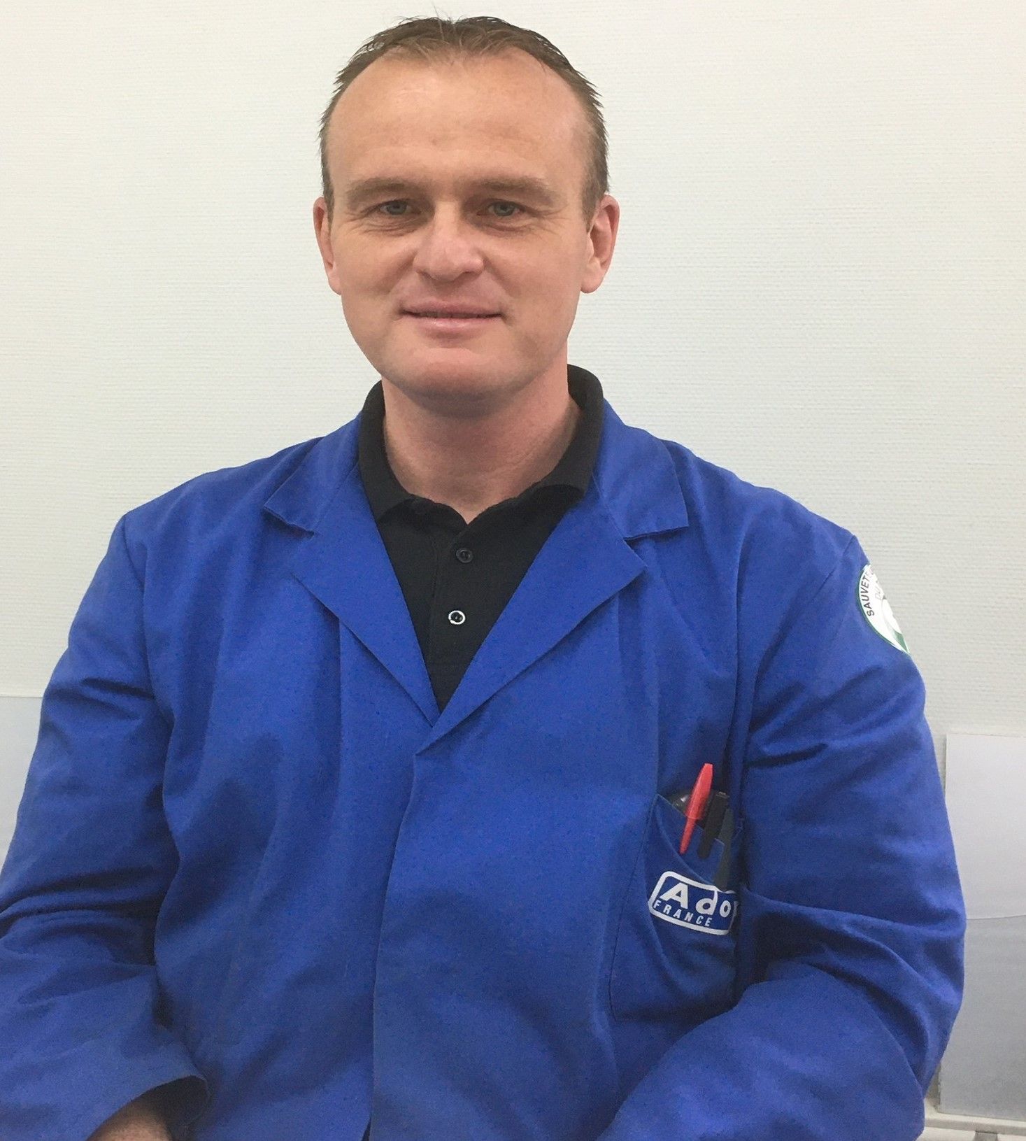 Un homme en blouse bleue de laboratoire se tient à l'intérieur, adossé à un mur clair.