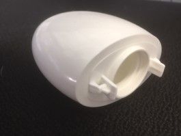 Couvercle de batterie en plastique blanc avec un petit loquet, posé sur une surface texturée sombre.