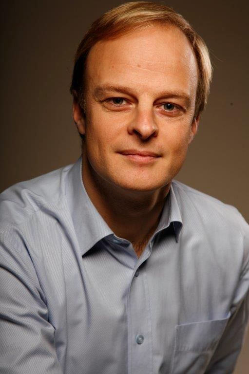 Portrait d'un homme en chemise bleu clair, de face, esquissant un léger sourire sur fond sombre.