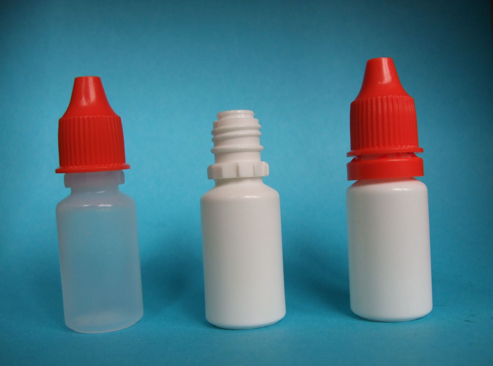 Trois petits flacons compte-gouttes en plastique sur fond bleu, deux avec des bouchons rouges et un flacon blanc.