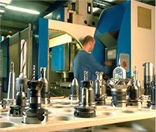 Ouvrier en chemise bleue manipulant une machine CNC dans une usine, avec des pièces métalliques sur un établi au premier plan.