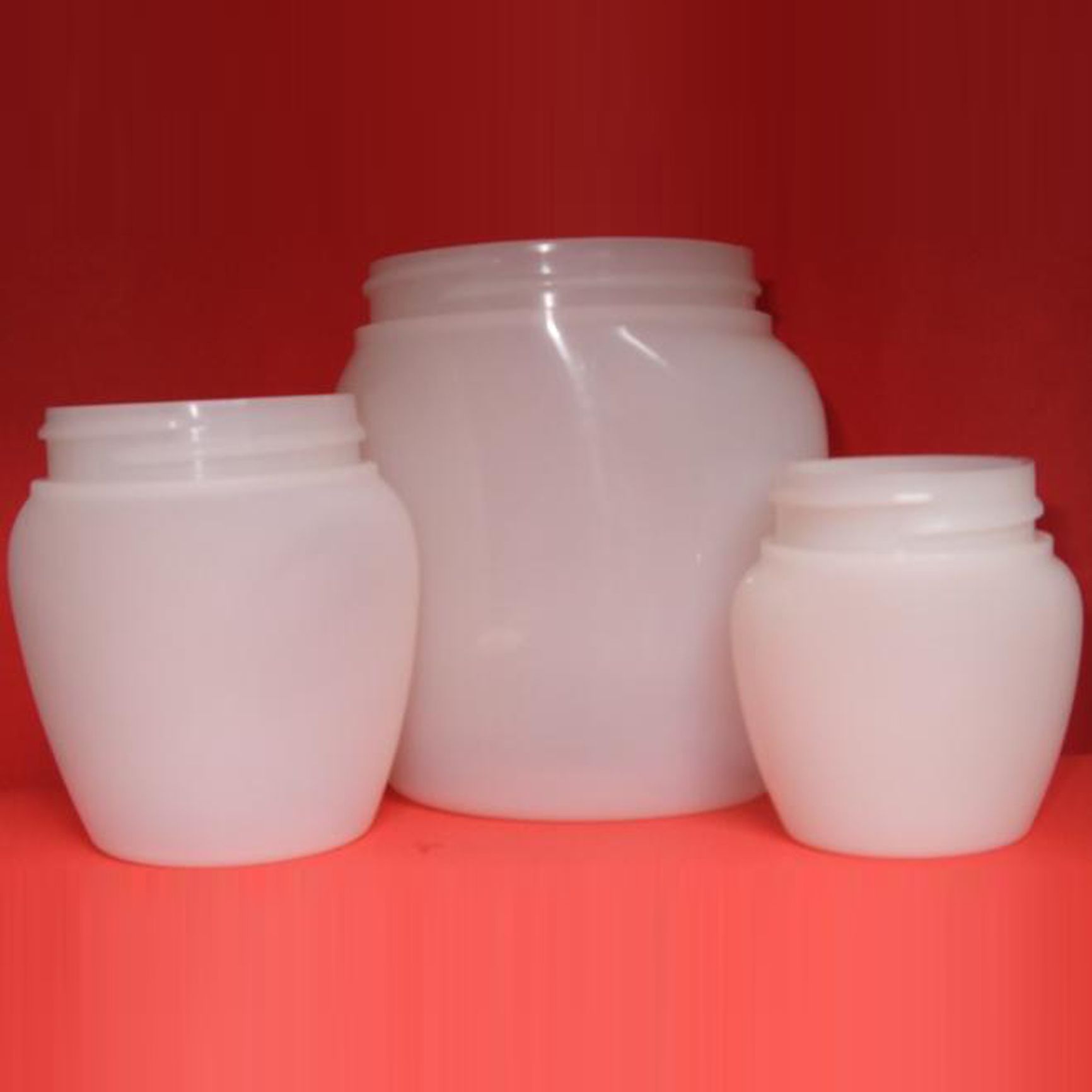 Trois bocaux en plastique blanc sur fond rouge, un grand bocal étant placé au centre, entre deux plus petits.