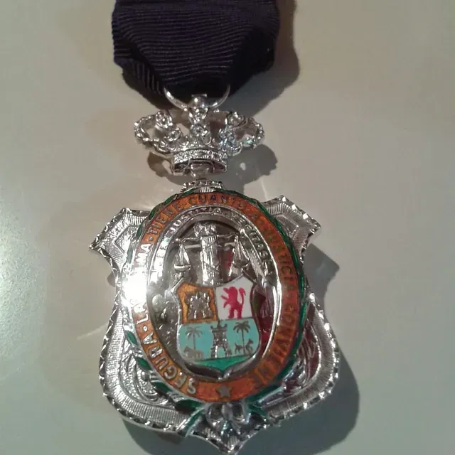 Medalla de plata con corona, escudo y cinta. El escudo contiene un escudo de armas dentro de un óvalo naranja.