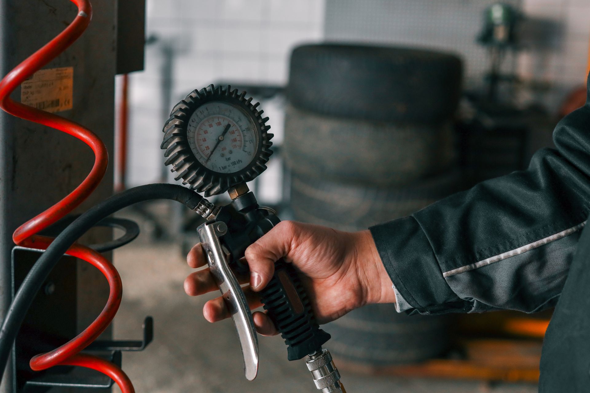 Une personne tient un manomètre de pression de pneus dans un garage ; un tuyau d’air rouge se trouve à proximité, des pneus sont visibles en arrière-plan.