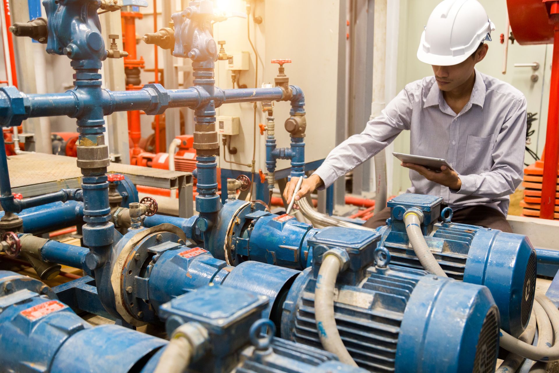 Un homme portant un casque de chantier inspecte des machines dans un environnement industriel, une tablette à la main.