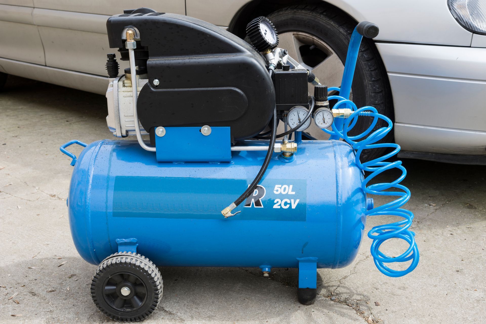 Compresseur d'air bleu et noir avec manomètres, tuyau et roues, près d'une voiture.