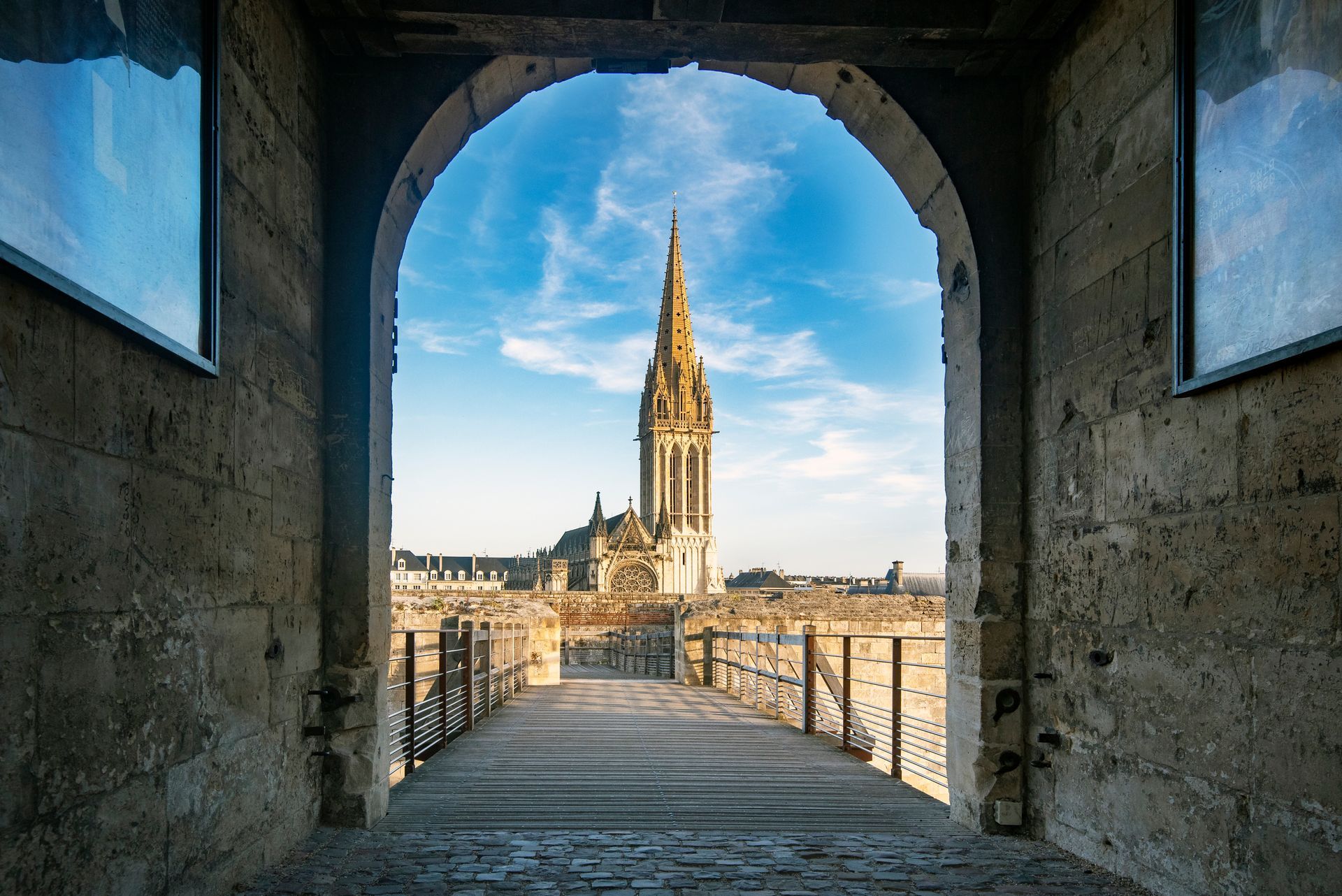 Une arche en pierre encadre la vue d'une haute flèche de cathédrale sous un ciel bleu.