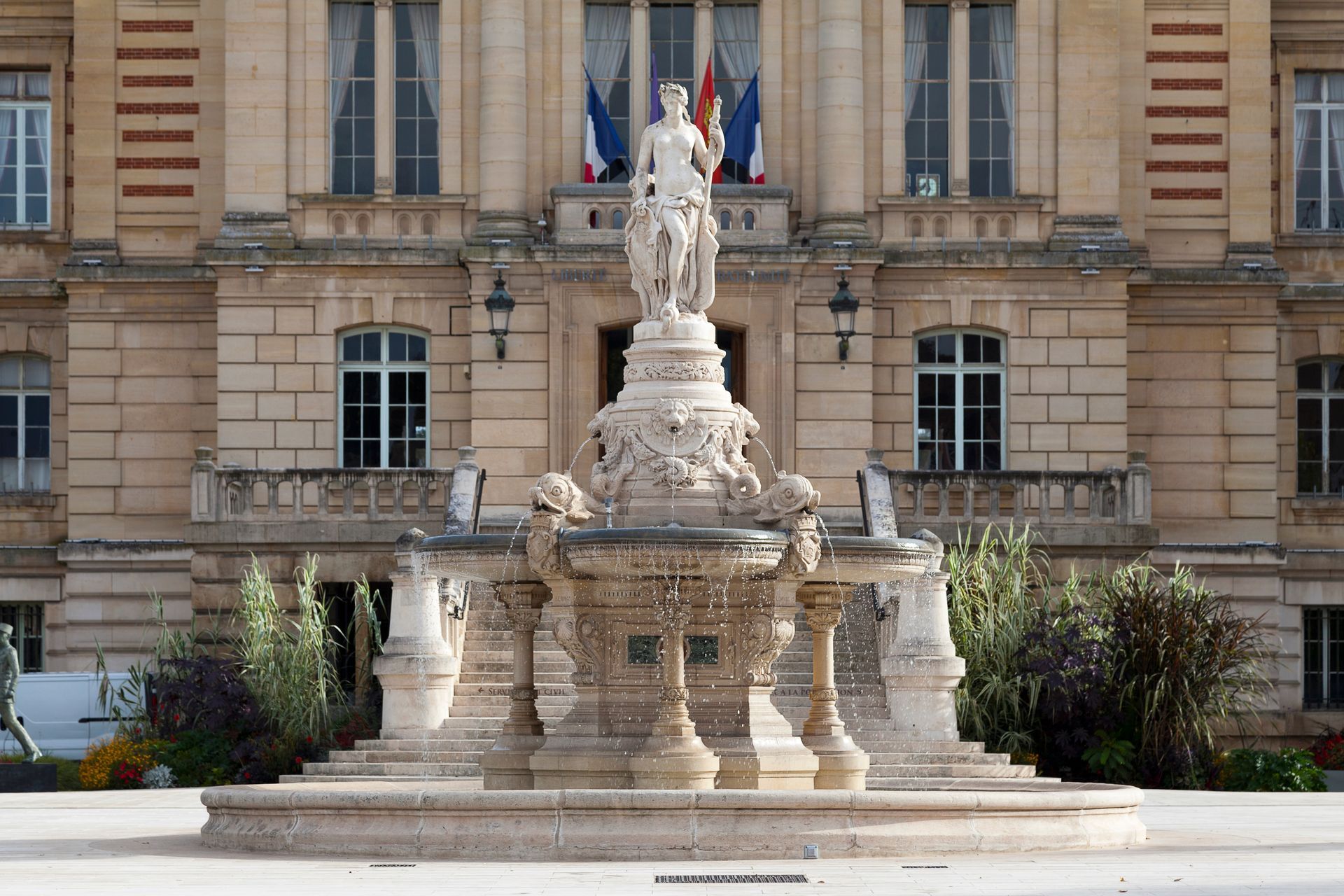 Fontaine ornée d'une statue devant un bâtiment en pierre pavoisé de drapeaux français.