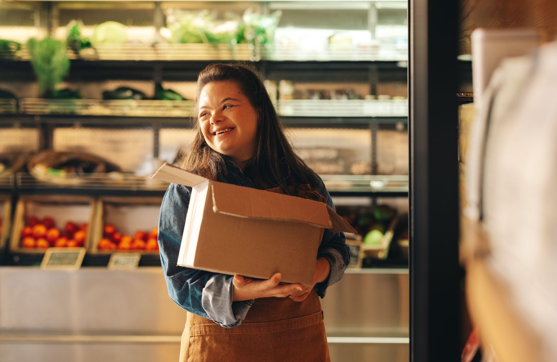 Personne travaillant en magasin tenant une boîte devant un étal de fruits et légumes