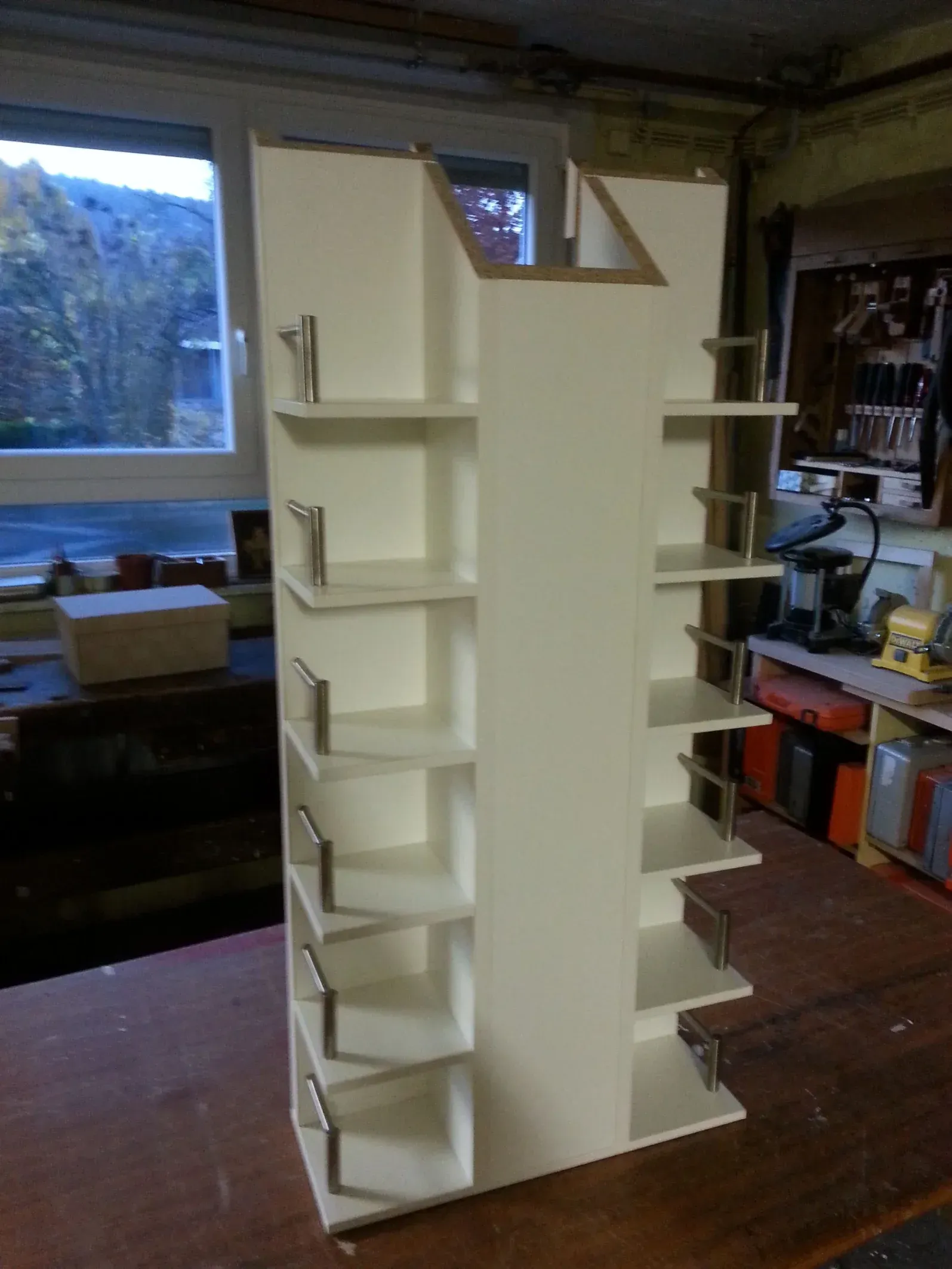 Ein hohes, weißes, drehbares Bücherregal mit mehreren Ablagen und silbernen Stützen steht auf einer Holzfläche in der Nähe eines Fensters.