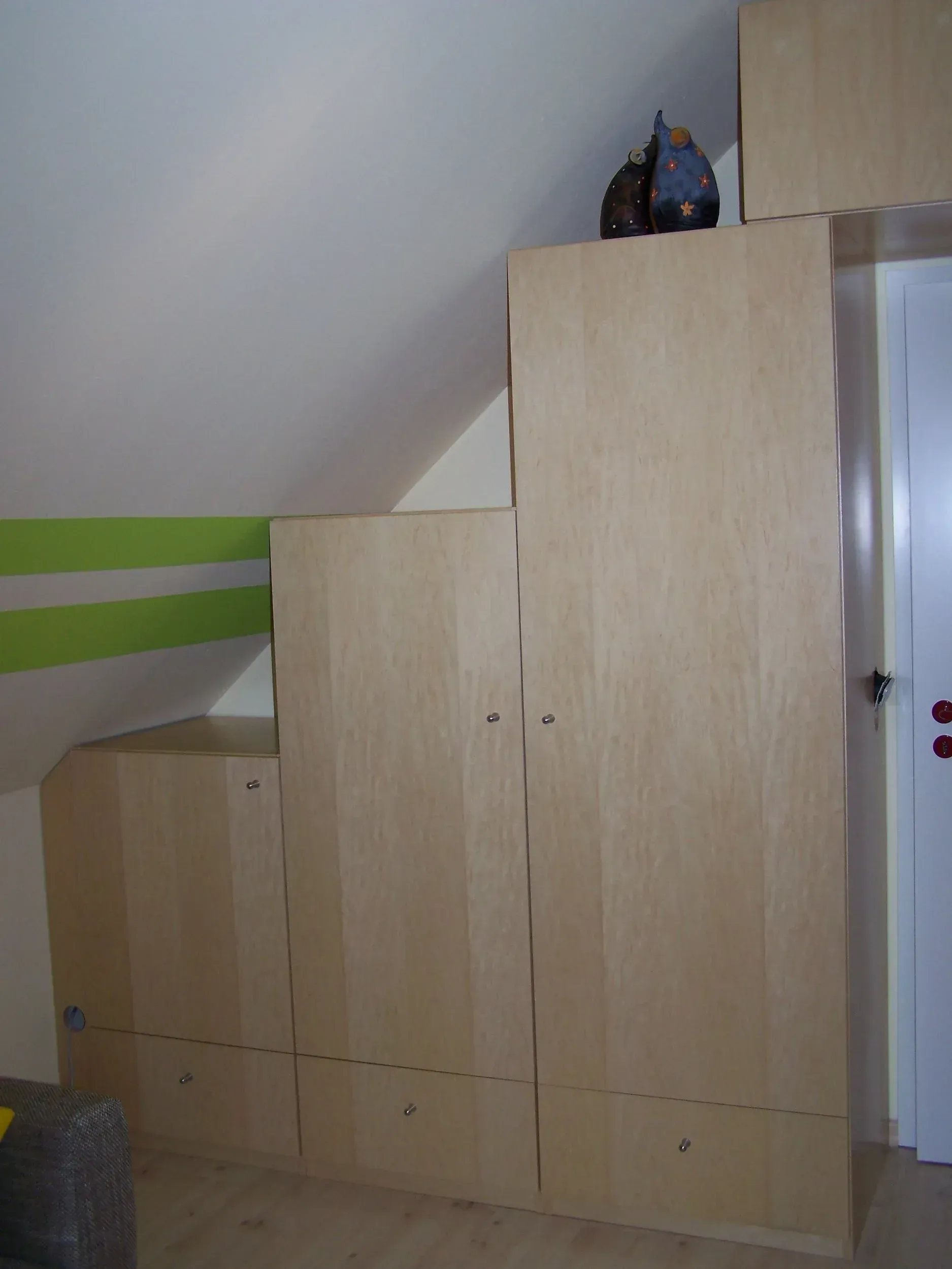 Eingebaute Holzschränke in einem Raum mit Dachschräge; grüne Streifen an der Wand.