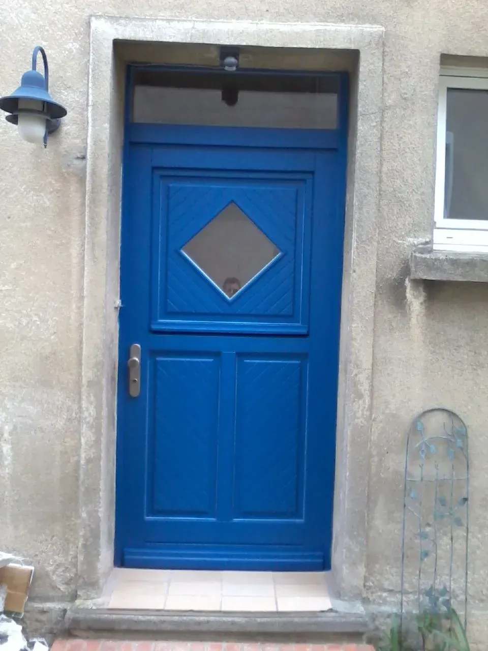 Blaue Eingangstür mit rautenförmigem Fenster und Seitenfenster. Sie befindet sich in einem Steingebäude mit einer Laterne und einem bogenförmigen Gartenstecker.