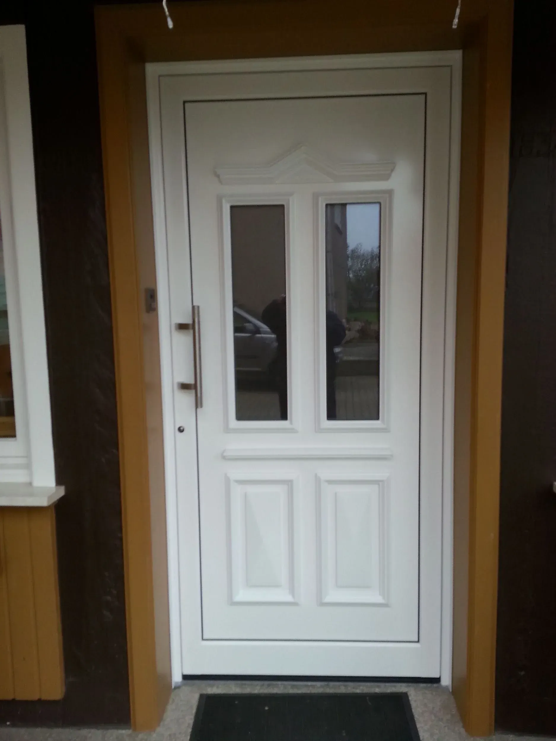 Weiße Haustür mit zwei Fensterscheiben und einem langen silbernen Türgriff. Braune Zierleisten.