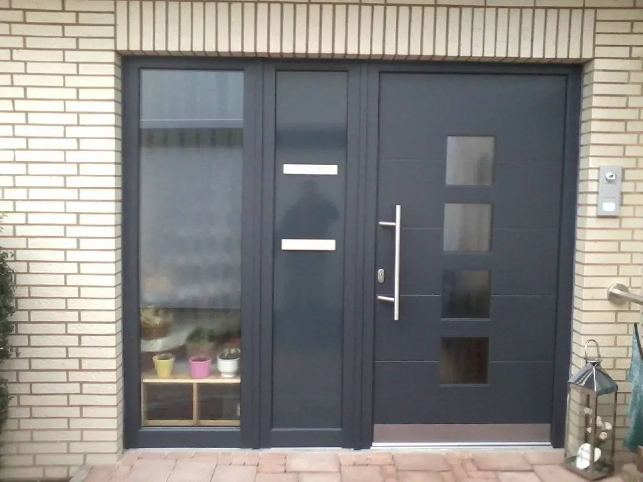 Dunkelgraue Eingangstür mit Seitenfenster und Glaseinsätzen, in Ziegelstein eingelassen.