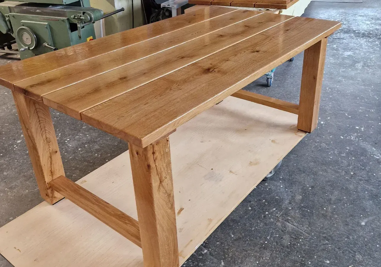 Holztisch mit hellbrauner, plankenartiger Tischplatte und stabilen Beinen, der auf einer Arbeitsfläche steht.