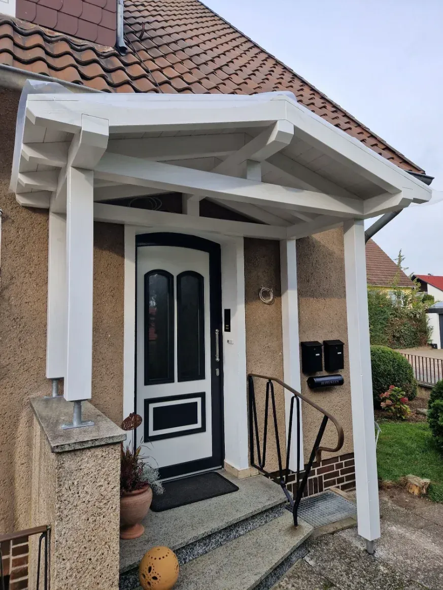 Veranda mit weißem Rahmen und schwarzer Tür. Geschwungenes Geländer, Briefkasten und Topfpflanze sind zu sehen.