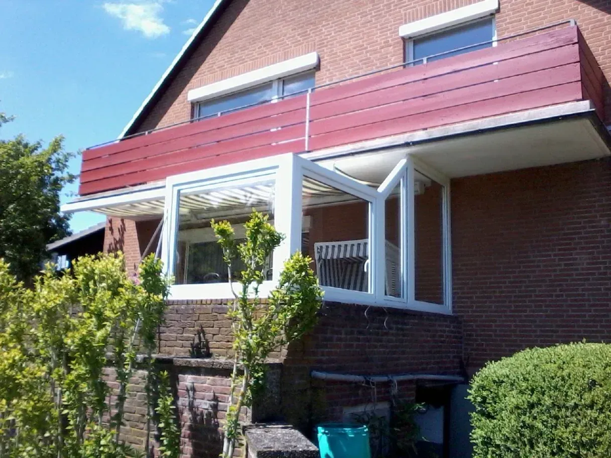Ein Backsteinhaus mit einem rotgedeckten Balkon und einem Wintergarten mit großen Fenstern und weißem Rahmen.