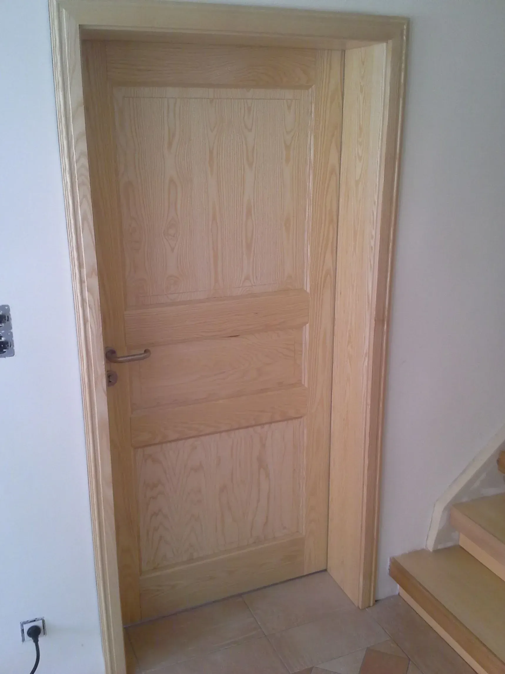 Holztür in hellem Holzrahmen, halb geöffnet. Treppe rechts, helle Wände und Zierleisten.