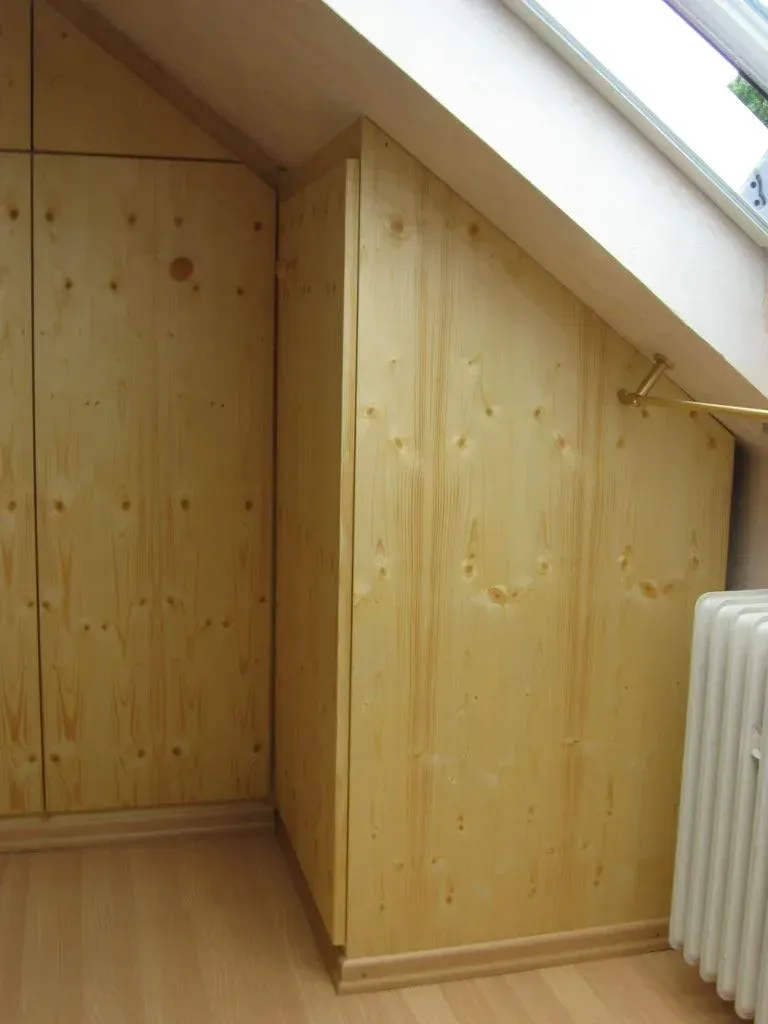 In einem Zimmer mit Dachschräge und Fenster befindet sich ein hölzerner Einbauschrank. Laminatboden und ein Heizkörper sind ebenfalls zu sehen.