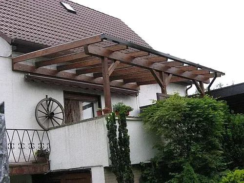 Eine hölzerne Pergola über einem Balkon an einem weißen Haus mit dunkelbraunem Dach, Pflanzen und einem antiken Rad.