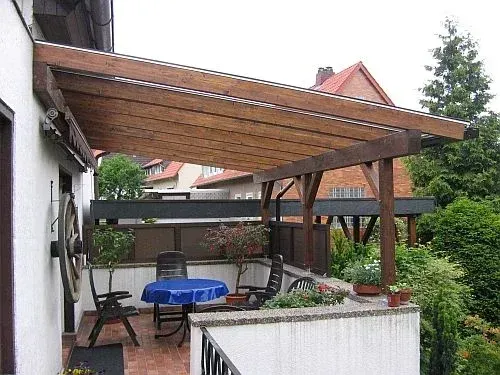 Eine hölzerne Pergola über einem Balkon mit Tisch und Stühlen bietet Blick auf einen Garten.