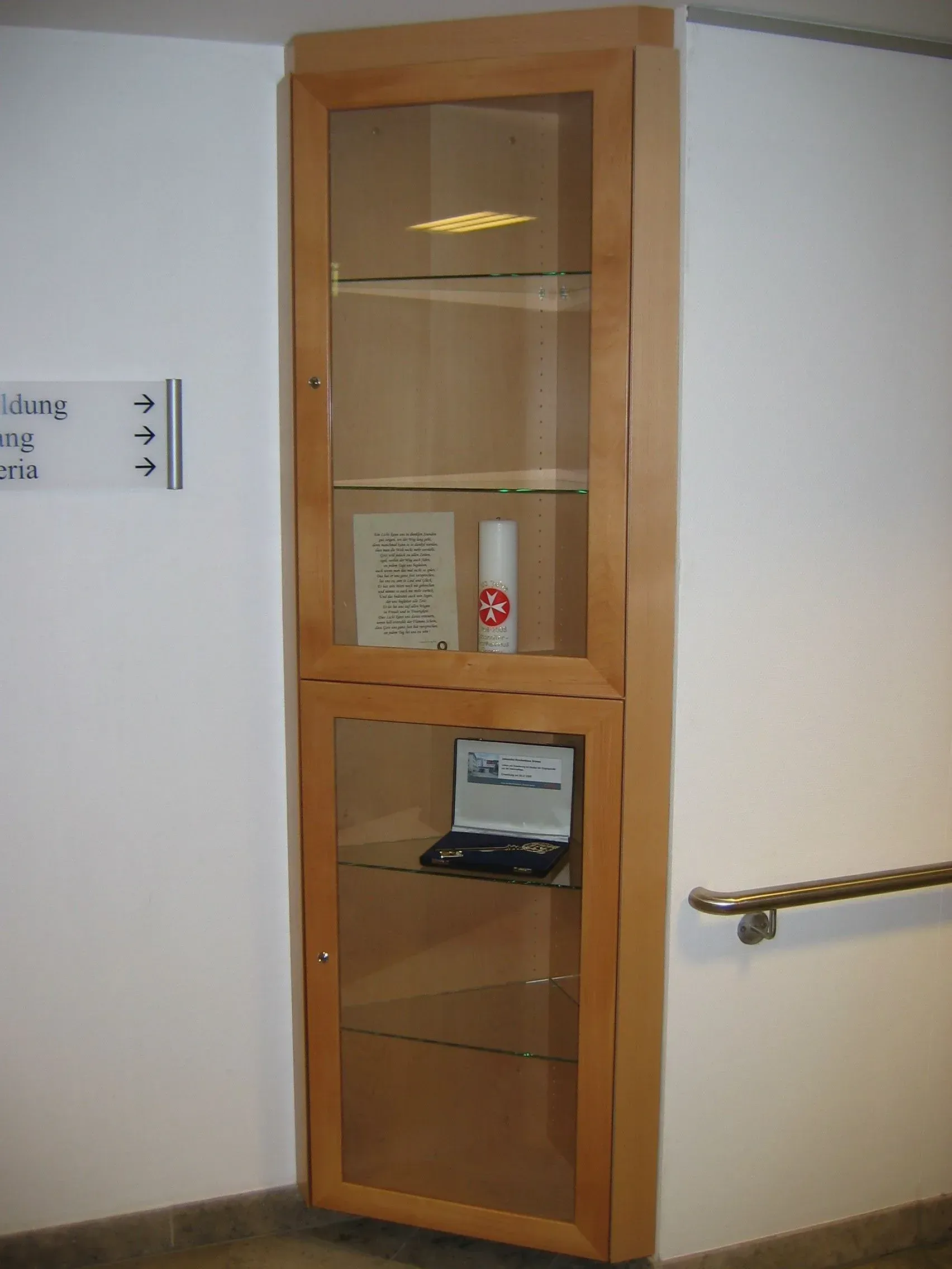 Eckvitrine mit Glastüren, die Gegenstände enthält, an einer weißen Wand in einem Flur angebracht.