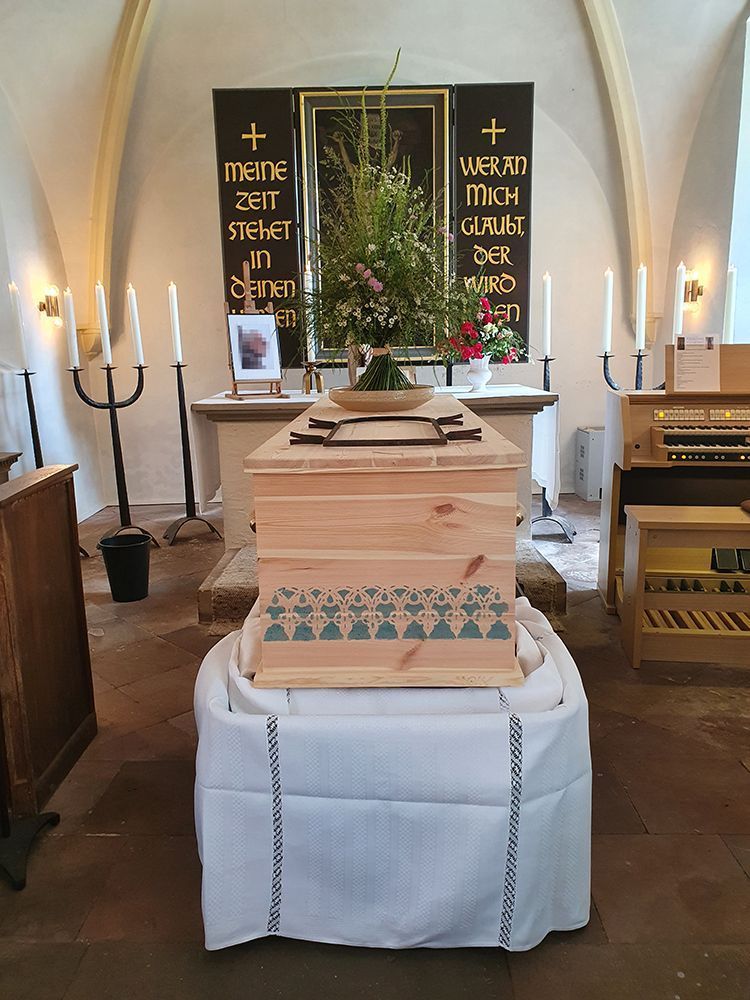 Ein Holzsarg auf einem drapierten Podest in einer Kirche. Blumen darauf. Kerzen und Orgel in der Nähe.