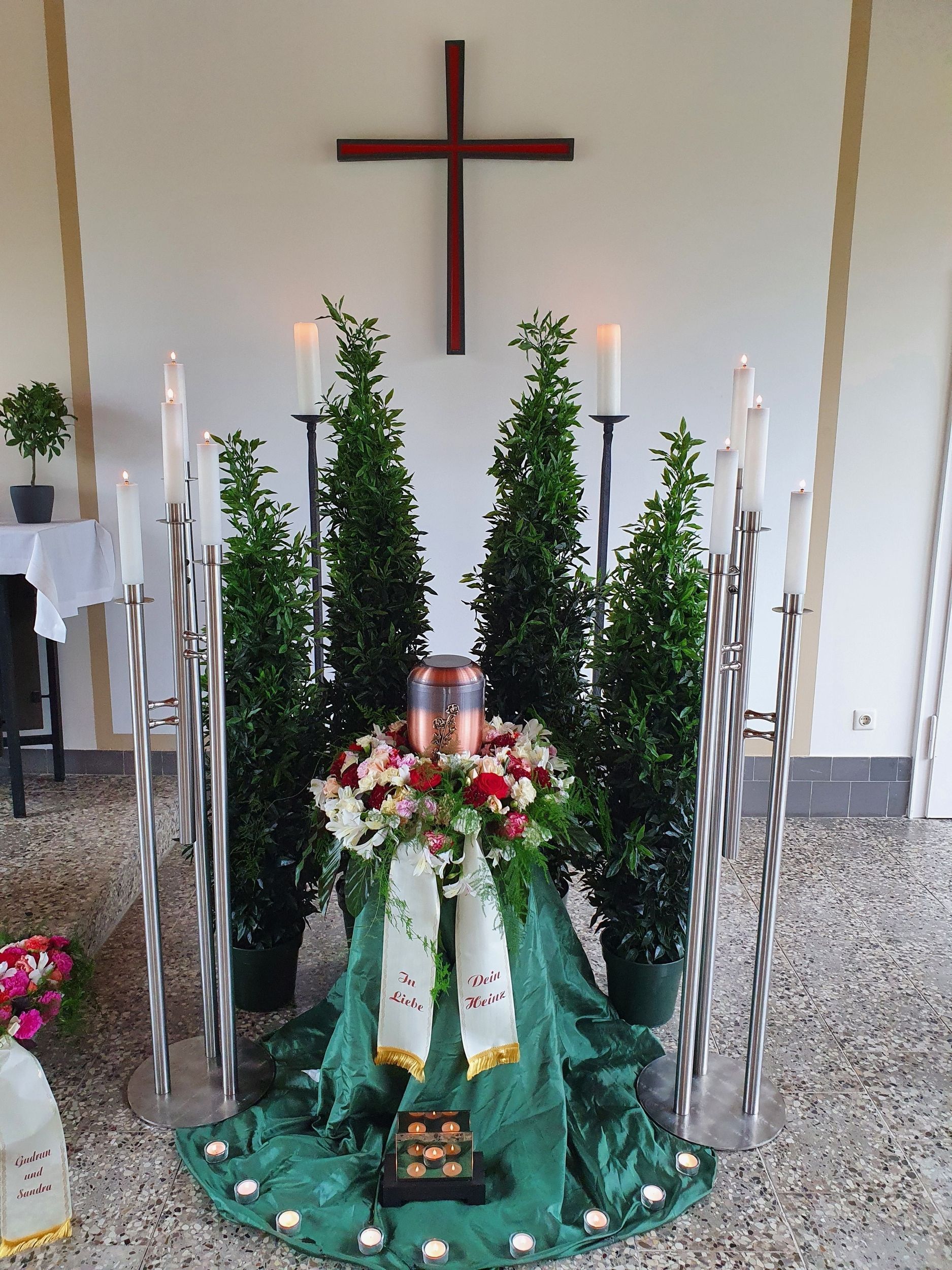 Trauerdekoration: Urne, Blumen, Kreuz, Kerzen. Grünes Tuch und Beiwerk.