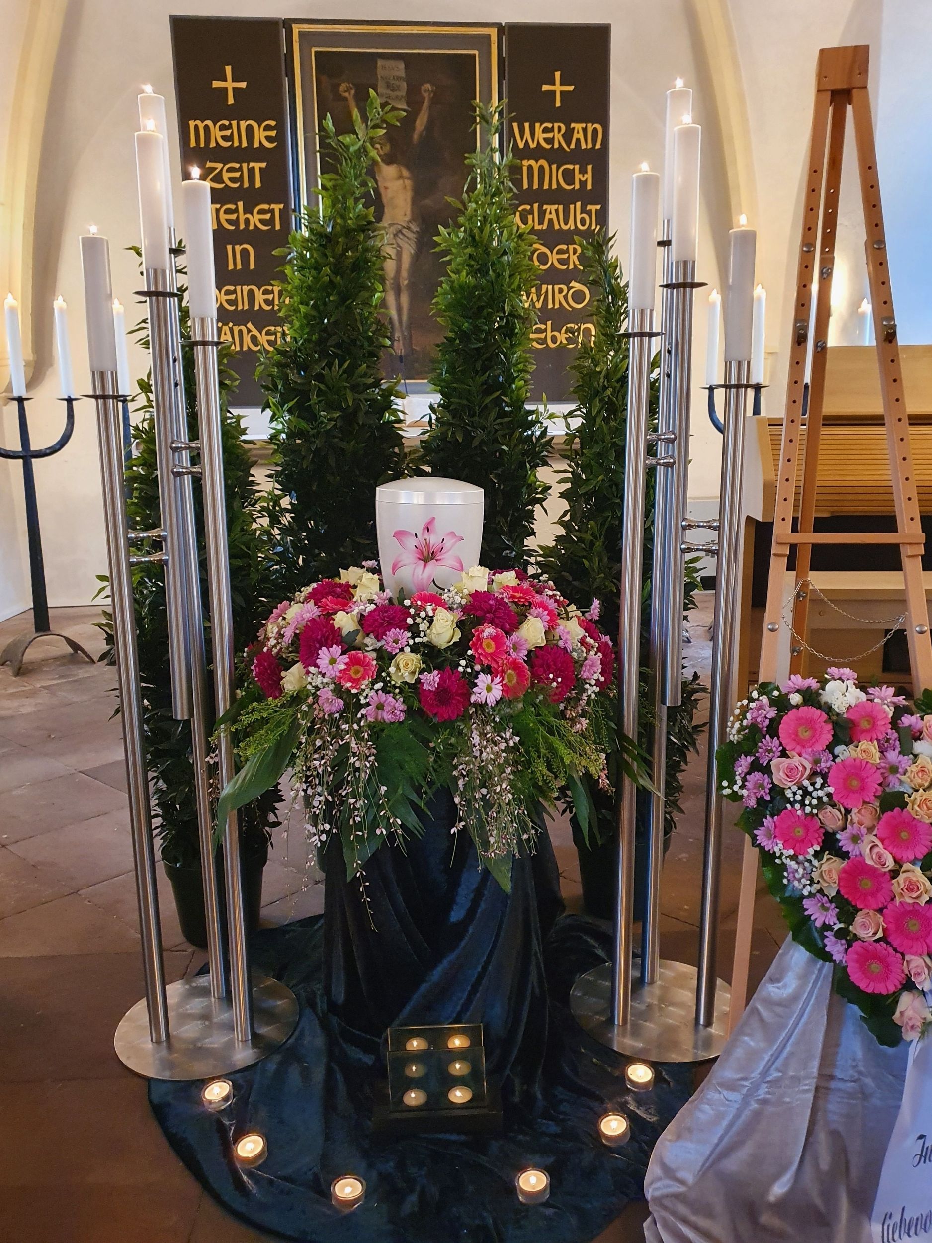 Trauerdekoration: Urne mit Blumenarrangement, Kerzen und immergrünen Zweigen. Dunkler Hintergrund mit Text.