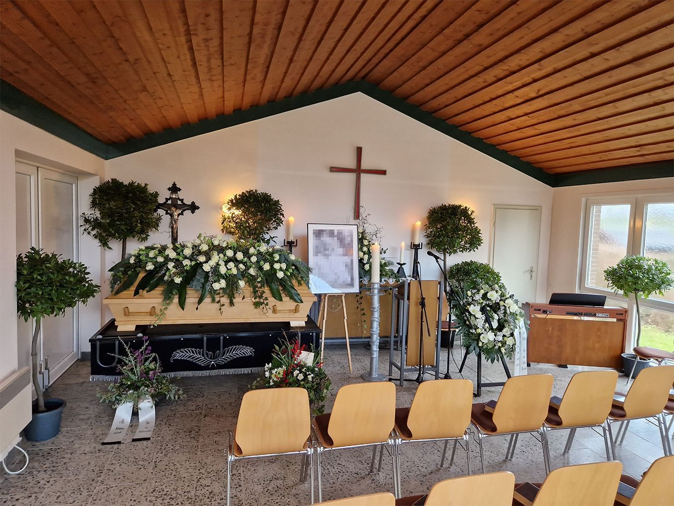 Trauerhalle mit Sarg, Blumen, Kreuz und Stühlen.