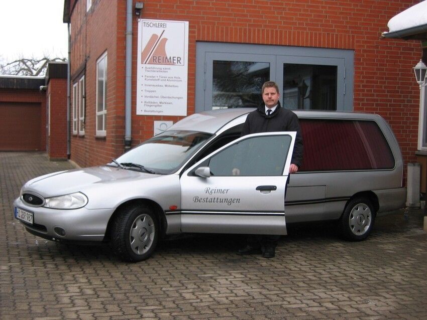 Ein Mann im schwarzen Anzug steht neben einem silbernen Leichenwagen mit offener Tür; im Hintergrund ist ein Gebäude zu sehen.
