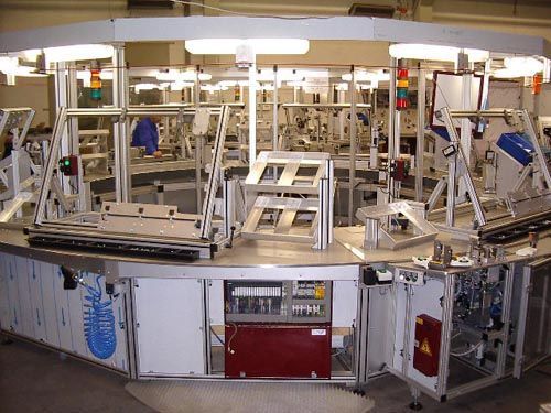Industrielle Montagestation mit kreisförmigem Fördersystem, Metallrahmen und Deckenbeleuchtung in einer Produktionsanlage.