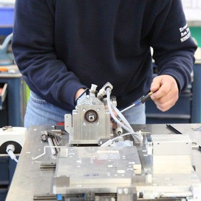 Ein Techniker montiert mit einem Schraubendreher ein mechanisches Metallbauteil auf einem Werkstatttisch.