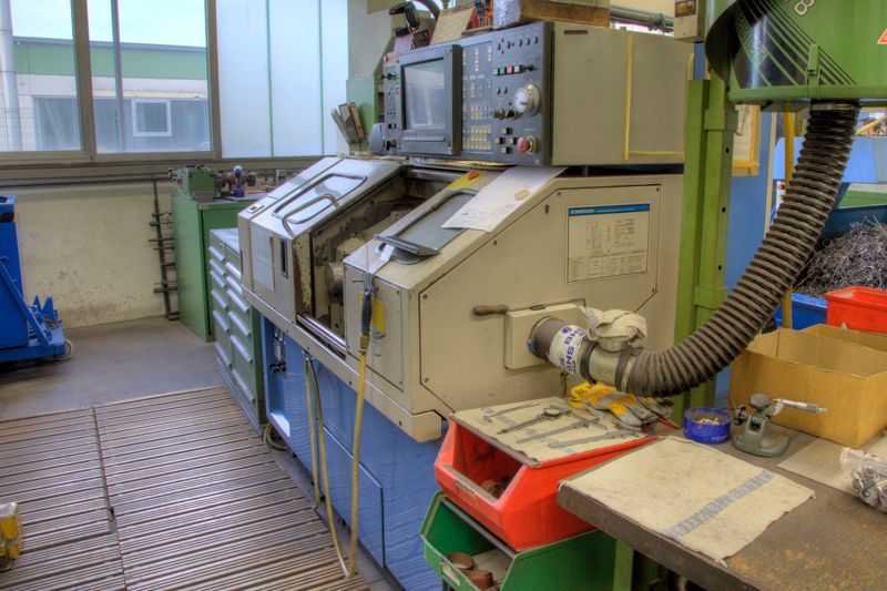 In einer Werkstatt steht eine CNC-Drehmaschine mit Bedienfeld neben einer Werkbank mit Werkzeugen und Aufbewahrungsbehältern.