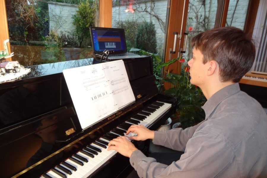 Un homme joue du piano noir, les yeux rivés sur une partition. Un ordinateur portable se trouve derrière le pupitre ; en arrière-plan, on aperçoit des fenêtres et de la verdure.