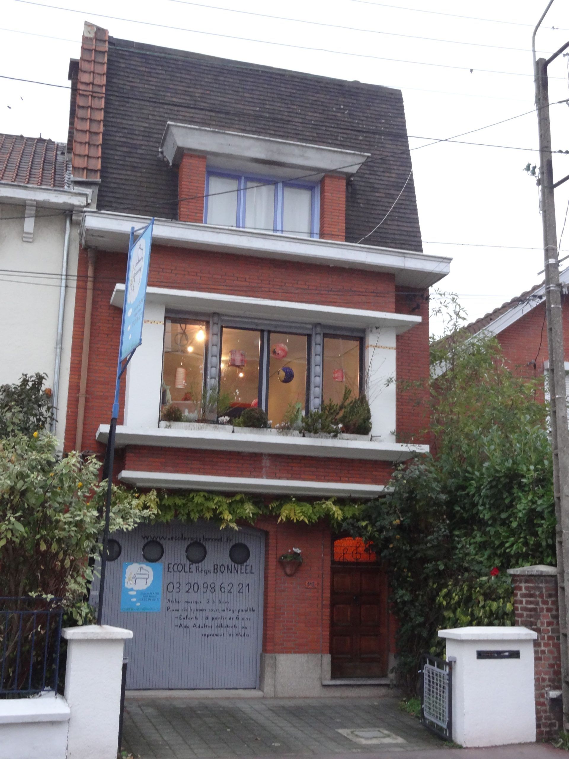 Bâtiment de deux étages en briques rouges avec garage, porte et fenêtres. Le dernier étage, au toit sombre, possède une petite fenêtre.