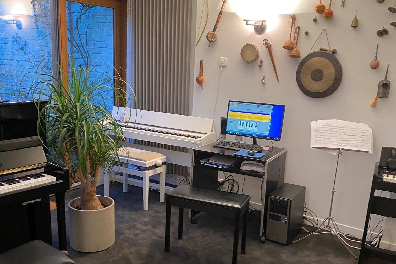 Une salle de musique avec un piano blanc, des instruments, un ordinateur et une grande plante.