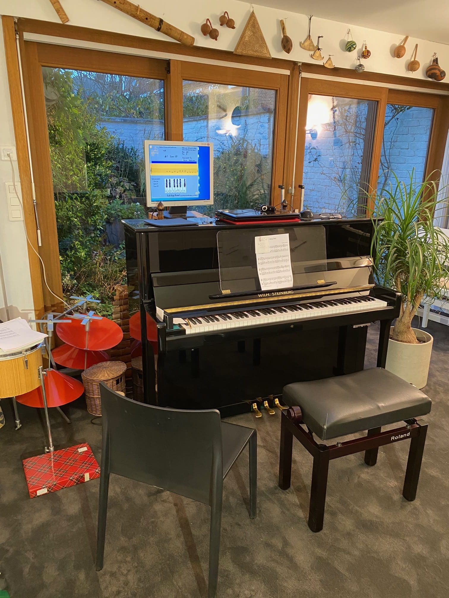 Piano noir près des fenêtres avec partitions, écran d'ordinateur, tabouret et chaise.