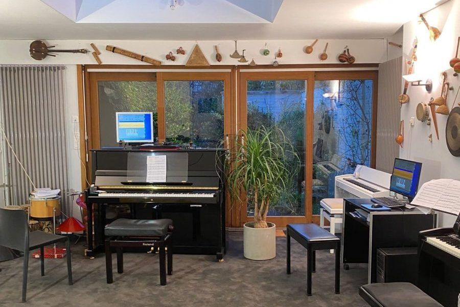 Salle de musique avec pianos, instruments accrochés aux murs et une grande plante. Les fenêtres donnent sur un jardin.