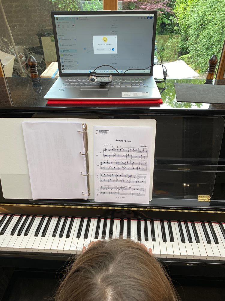 Une personne joue du piano, face à une partition, un ordinateur portable ouvert au-dessus des touches. La lumière du soleil entre par une fenêtre.