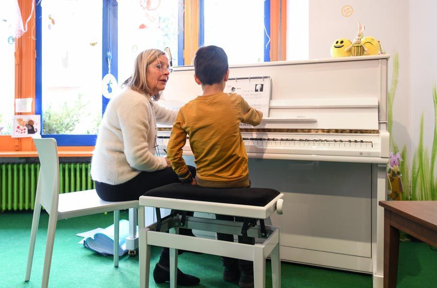 Une femme donne des instructions à un enfant au piano. Ils sont assis côte à côte ; les mains de l’enfant sont posées sur les touches. Pièce lumineuse.