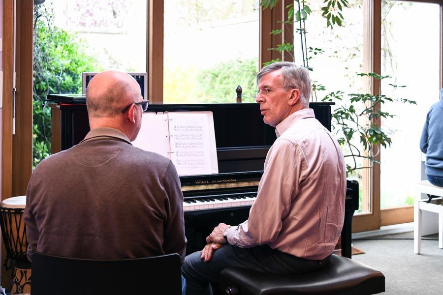 Deux hommes devant un piano : l’un face aux touches, l’autre en pleine conversation. Partitions sur le piano. Lumière naturelle éclatante.